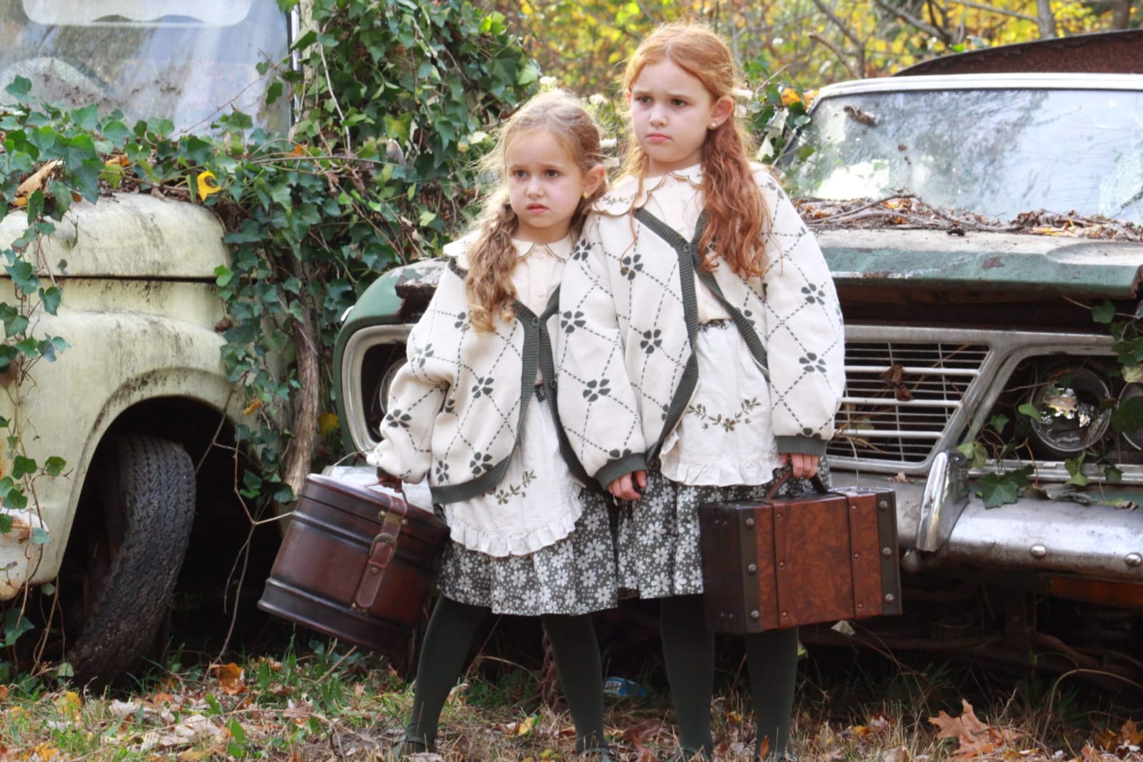 Two children holding suitcases beside vintage cars