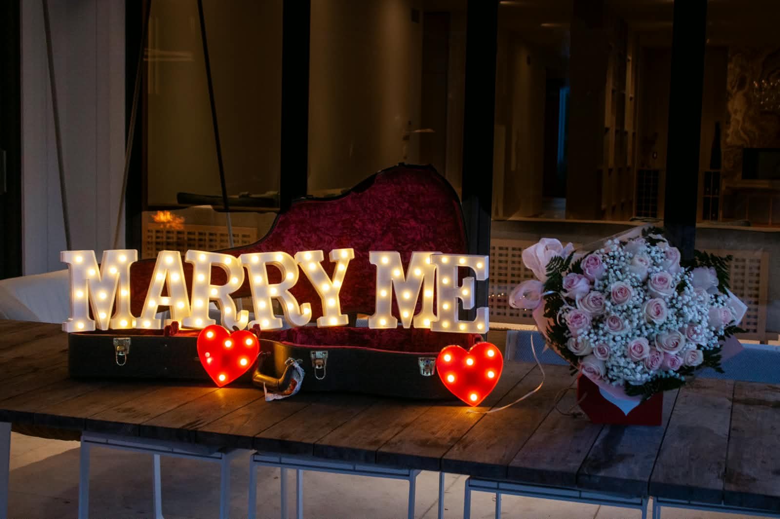 Illuminated MARRY ME letters in a guitar case with glowing red hearts and a pink rose bouquet