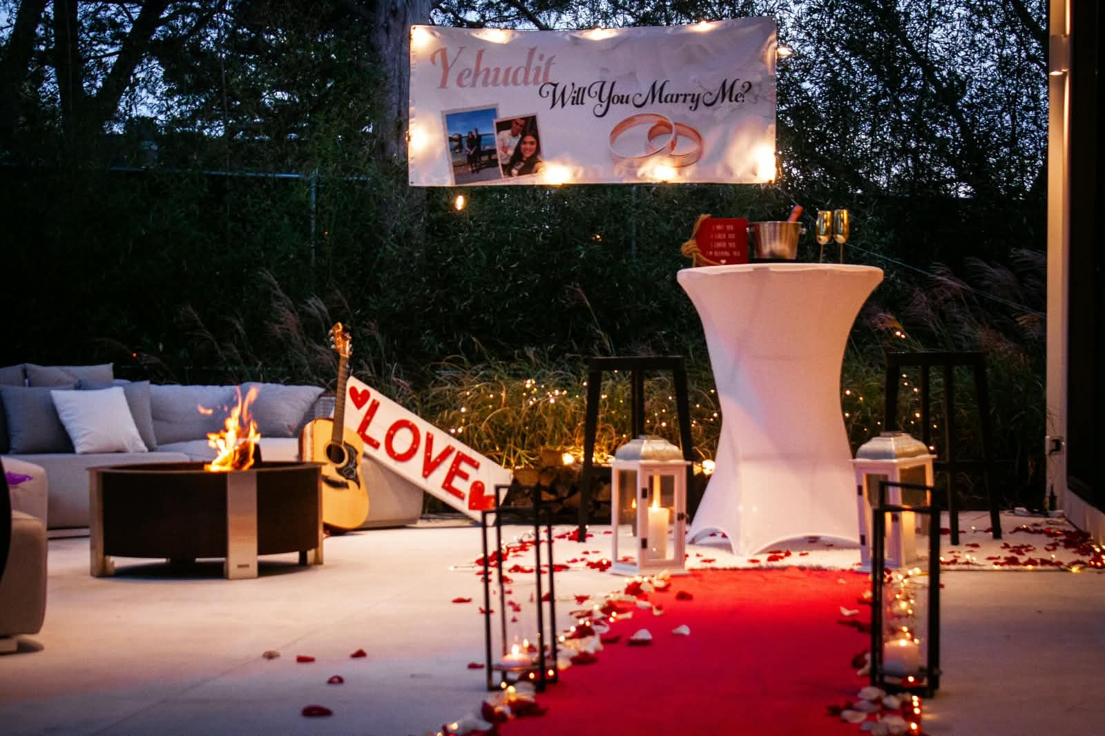 Outdoor evening proposal setup with red carpet, lanterns, LOVE sign and a Will You Marry Me? banner