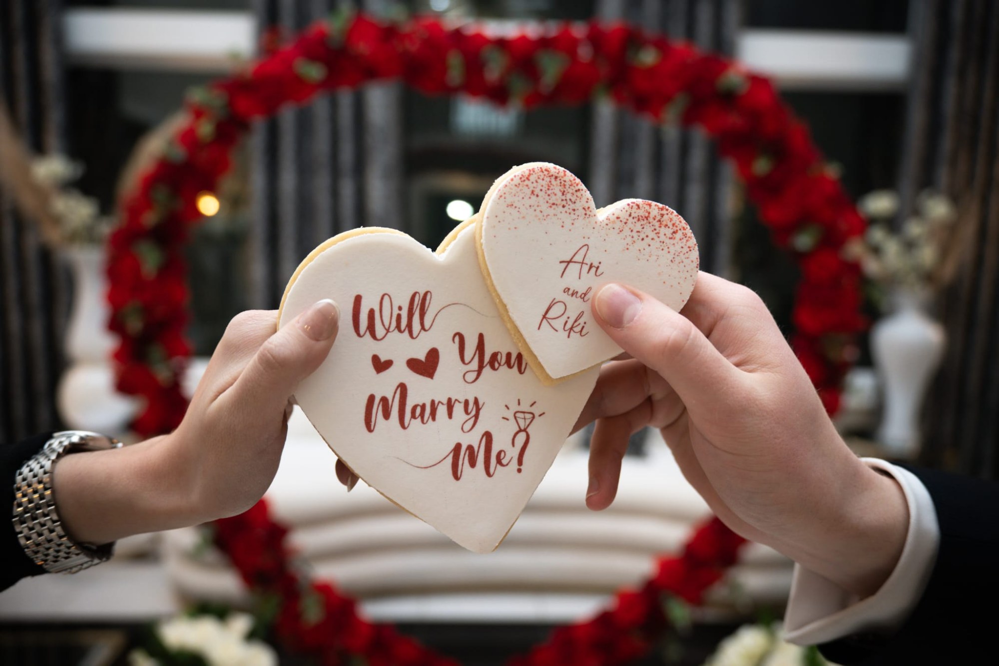 Hands holding heart-shaped Will You Marry Me? cookies in front of a red rose heart wreath